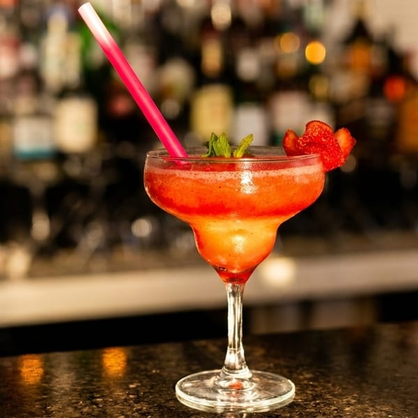 A strawberry margarita with a pink straw and mint garnish sits on a dark bar counter.