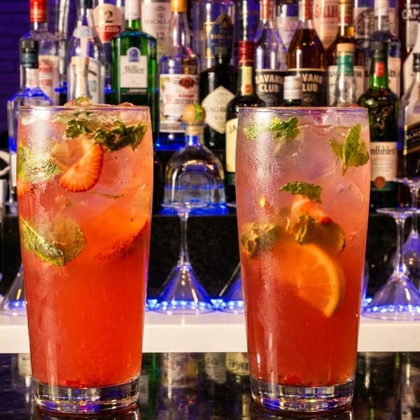 Two strawberry mojitos with mint and lime on a bar counter with liquor bottles in the background.