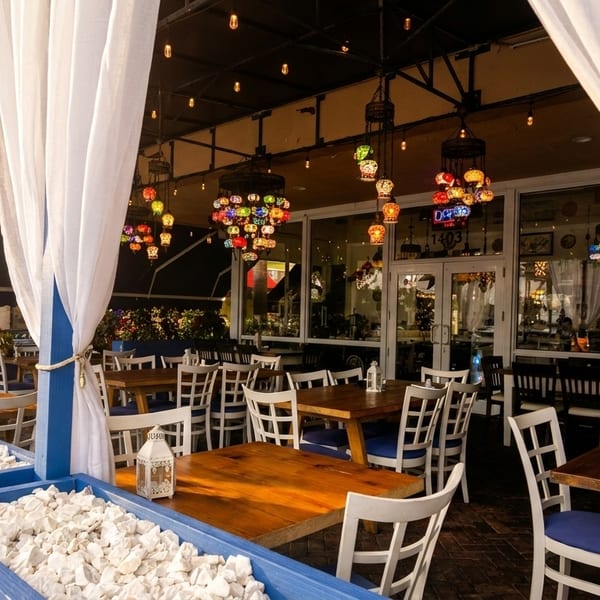 Outdoor patio dining area with white curtains, wooden tables, white chairs, and colorful hanging lamps.