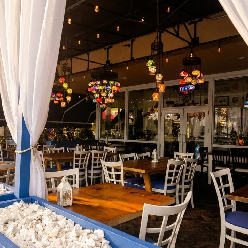 Outdoor patio dining area with white curtains, wooden tables, white chairs, and colorful hanging lamps.