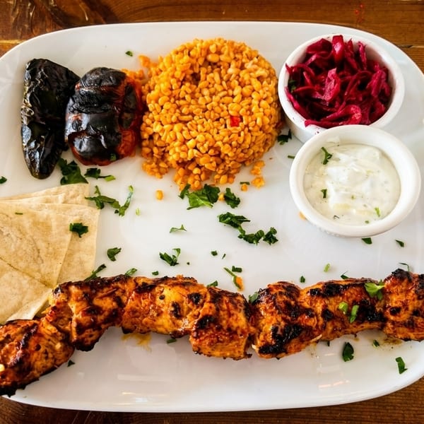 A white plate with chicken shish kebab, bulgur, grilled vegetables, flatbread, and dips on a wooden table.