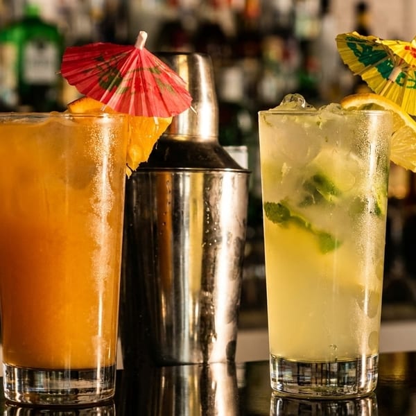 Two colorful cocktails with umbrellas and a shaker on a bar, with liquor bottles in the background.