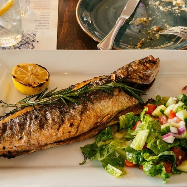 Grilled whole fish with rosemary, a lemon half, and a side salad on a white plate.