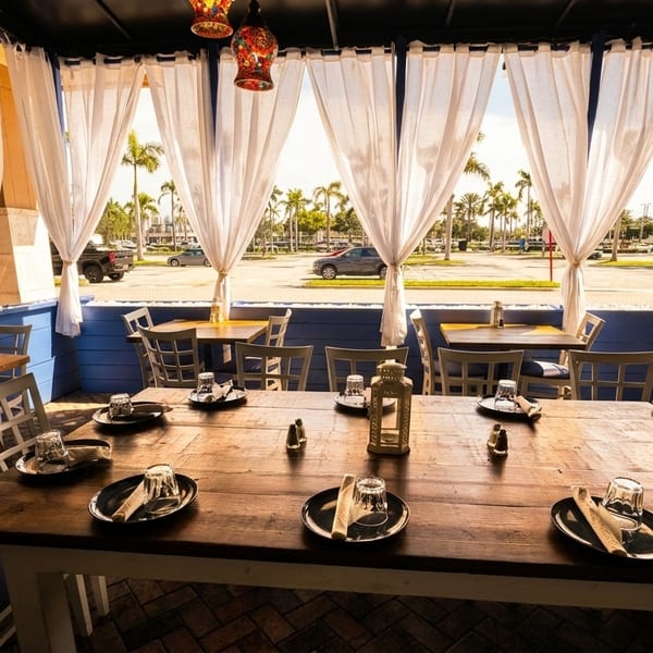 Outdoor restaurant patio with wooden tables, white chairs, and sheer curtains overlooking a street with palm trees.